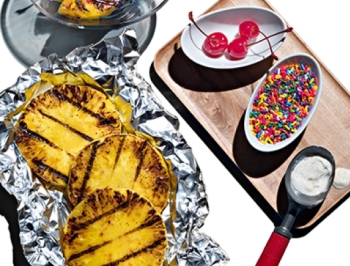 Grilled pineapple sitting on a sheet of aluminum foil alongside sundae toppings