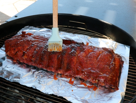 Person basting ribs sitting on an aluminum foil sheet on the grill with BBQ sauce