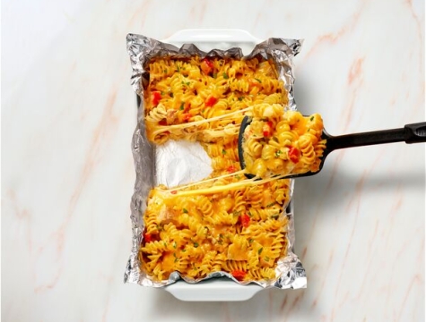 Person using a spatula to serve a cheesy pasta casserole from an aluminum foil lined baking dish